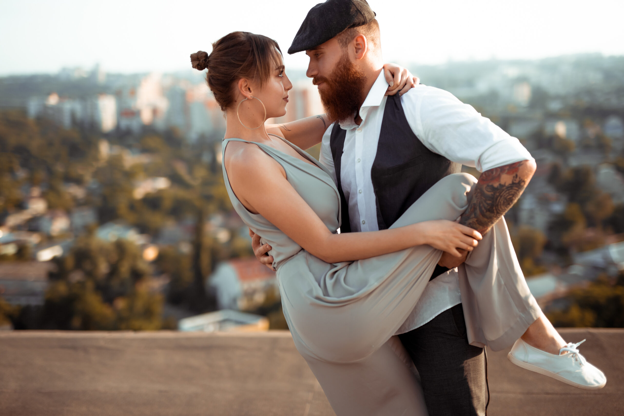 Trendy bearded hipster with tattooed arm hugging young girlfriend while dancing during romantic date on rooftop